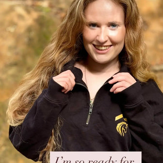 Person wearing a black jacket with an ABove Average equestrian logo, holding a sign saying 'I'm so ready for autumn', against a blurred natural background.