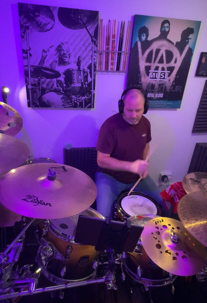 Above Average founder Adam Williams playing a drum kit in a home studio, wearing headphones and a dark tee, surrounded by cymbals and snare under purple lighting, with music posters on the wall — candid practice moment reflecting focus, presence, and the above average mindset rooted in creativity, sustainability, and everyday progress.